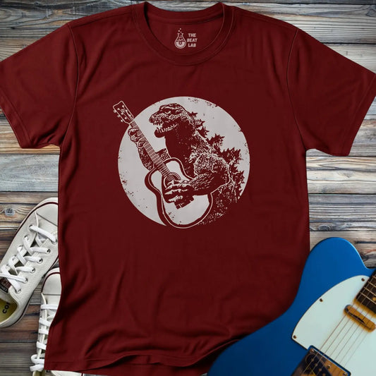 The Guitar Rex T-shirt is a maroon tee featuring an illustration of Godzilla playing an acoustic guitar within a circular backdrop, displayed on wooden boards beside white sneakers and a blue electric guitar.
