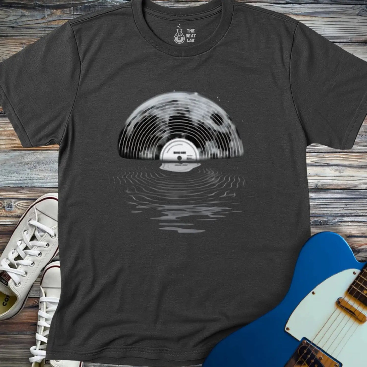 The Moon Vinyl T-shirt features a vinyl record half-submerged in water, creating a unique reflection. Displayed on a wooden surface beside white sneakers and a blue electric guitar.
