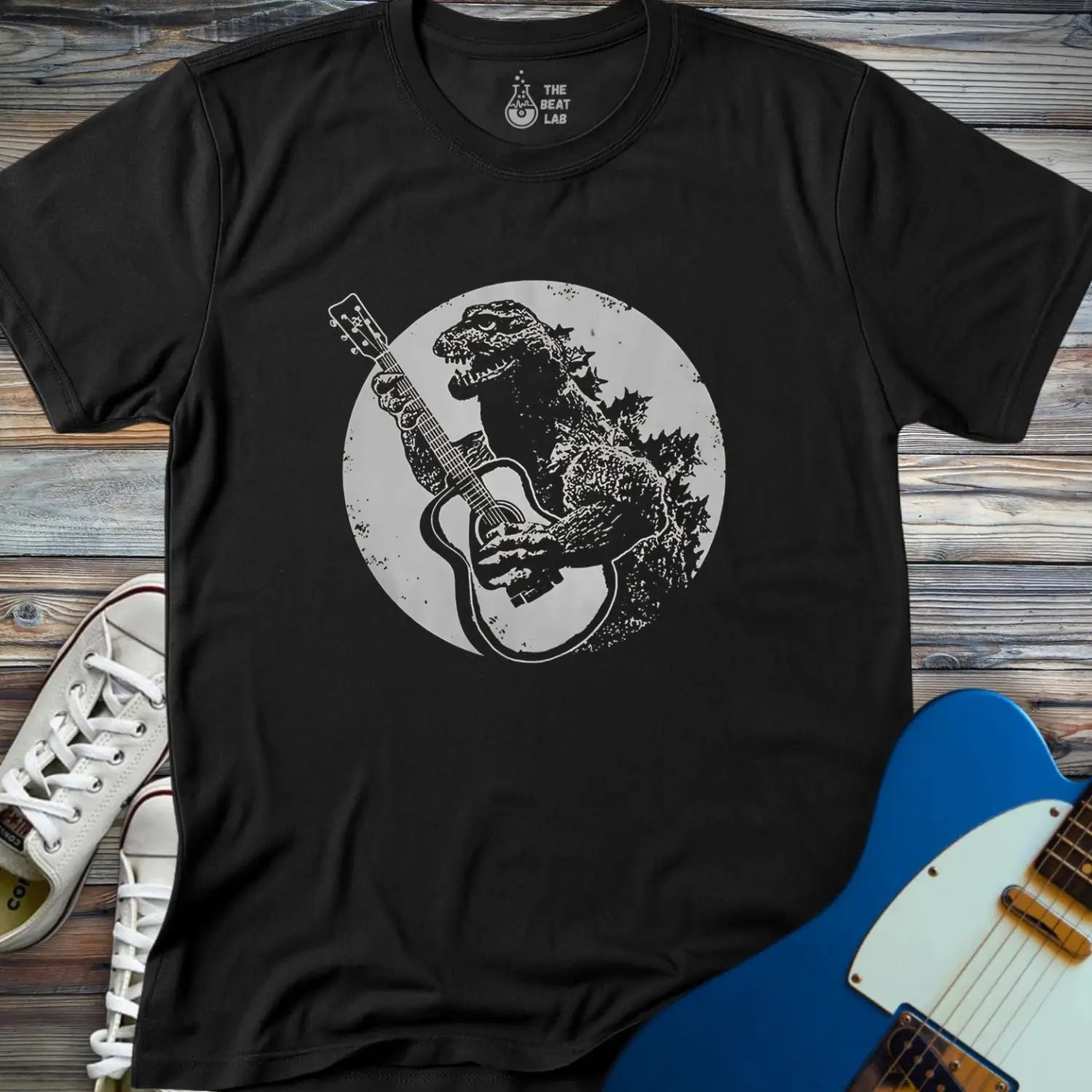 The Guitar Rex T-shirt is a black tee featuring Godzilla playing an acoustic guitar before a full moon, displayed on a wooden surface with white sneakers and a blue electric guitar.