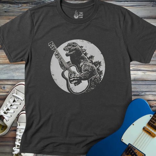 The Guitar Rex T-shirt features a charcoal gray design with Godzilla playing an acoustic guitar inside a circular background. Shown on wood beside white sneakers and a blue electric guitar.