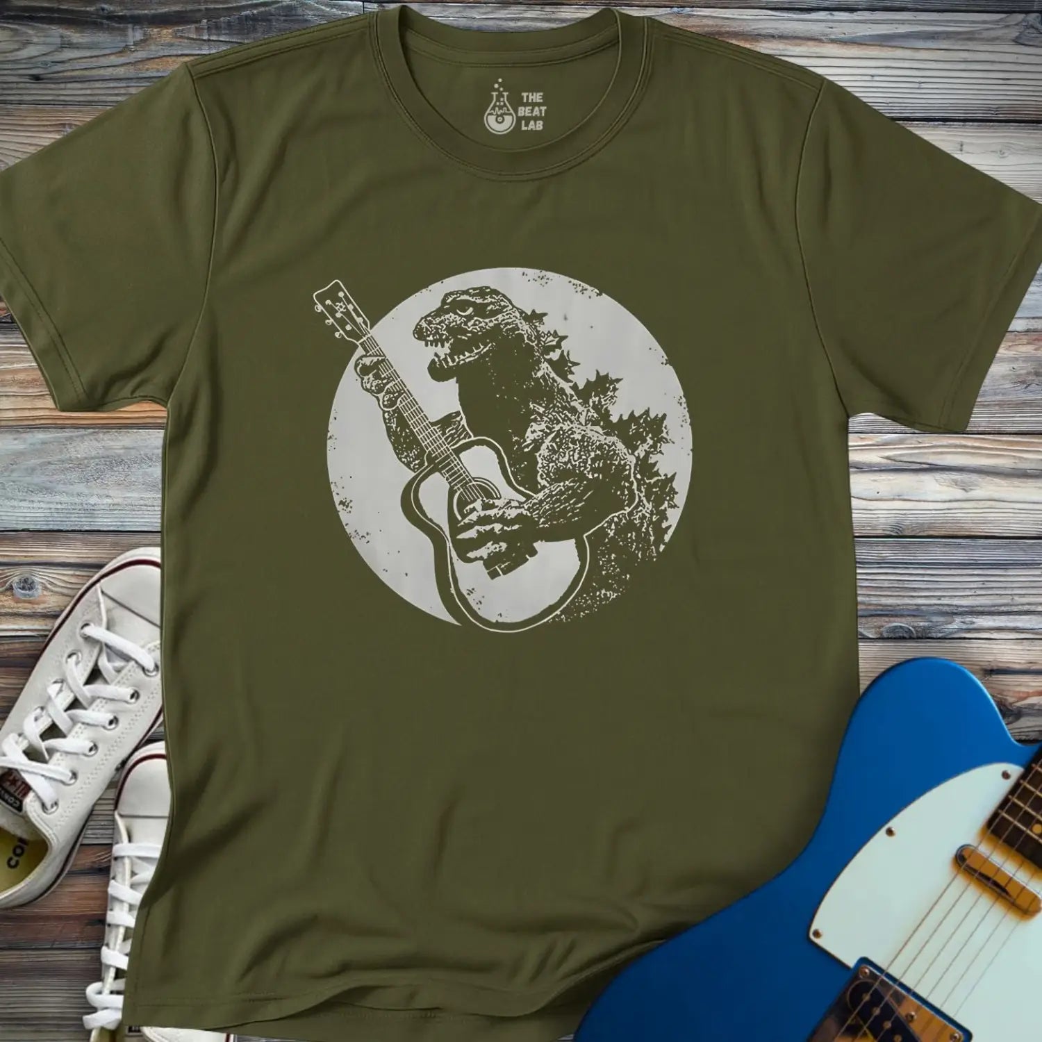 The Guitar Rex T-shirt is olive green and features Godzilla playing an acoustic guitar under a full moon. Shown laid on wood beside white sneakers and a blue electric guitar.
