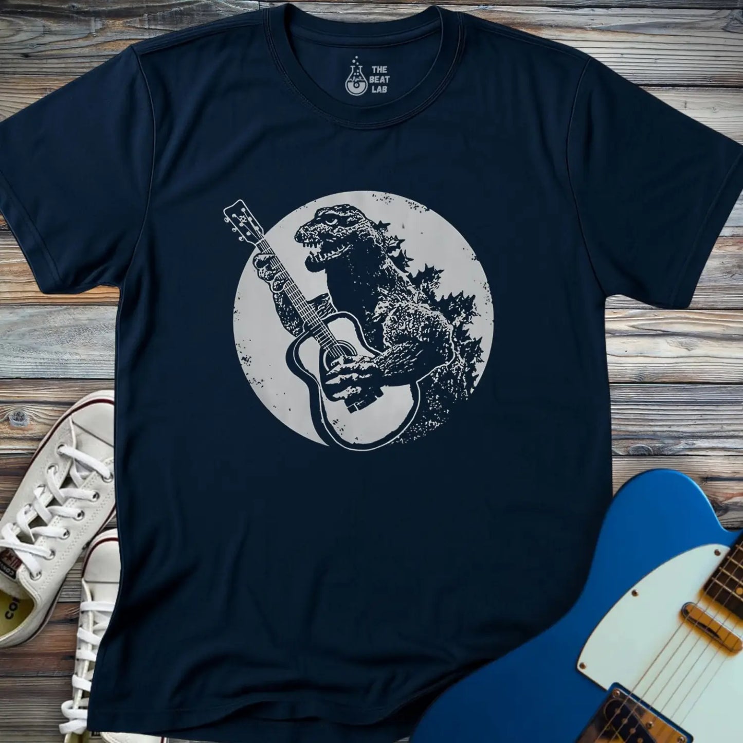 The Guitar Rex T-shirt is a navy blue tee with a dinosaur playing an acoustic guitar in front of a full moon, shown on a wooden surface alongside white sneakers and a blue electric guitar.