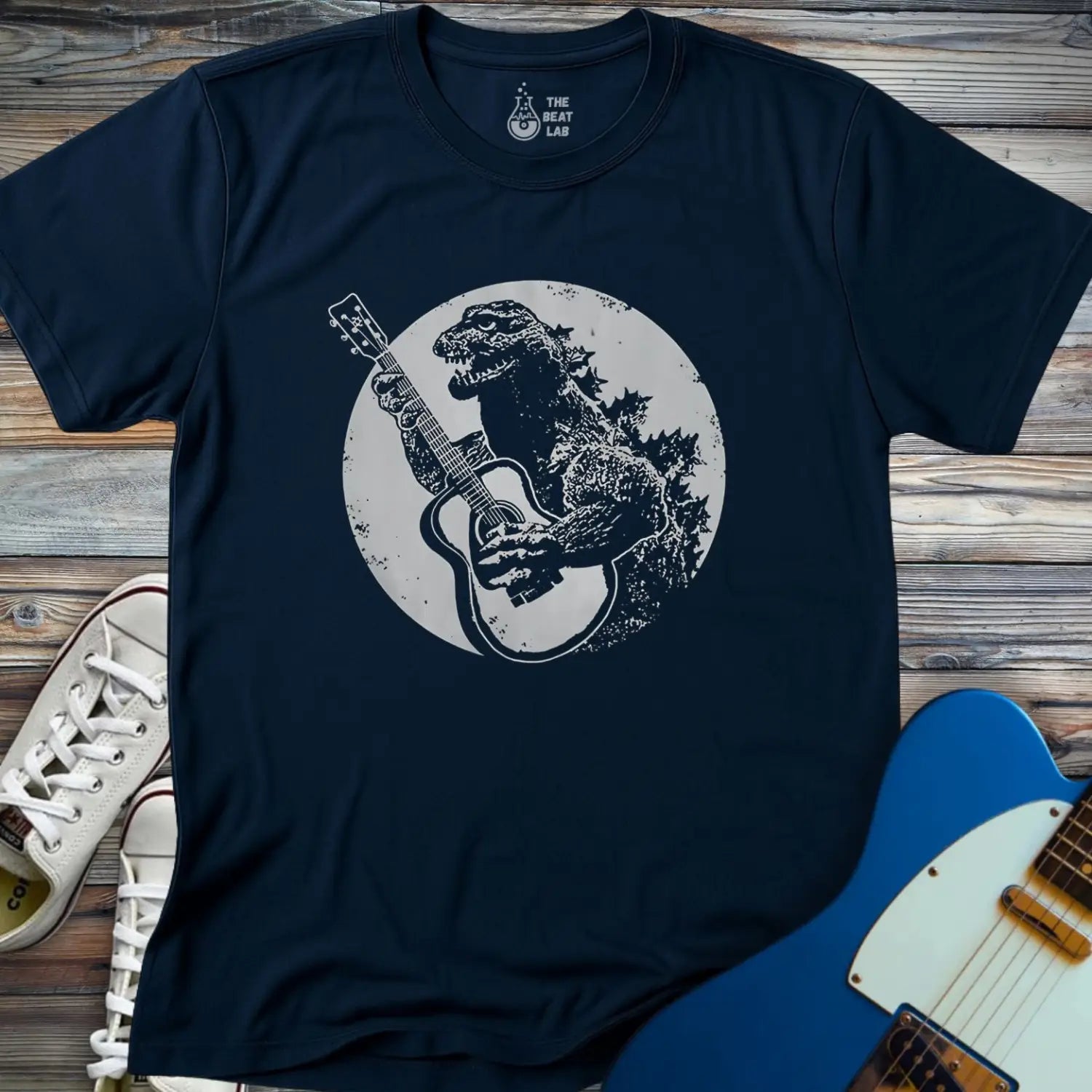 The Guitar Rex T-shirt is a navy blue tee with a dinosaur playing an acoustic guitar in front of a full moon, shown on a wooden surface alongside white sneakers and a blue electric guitar.