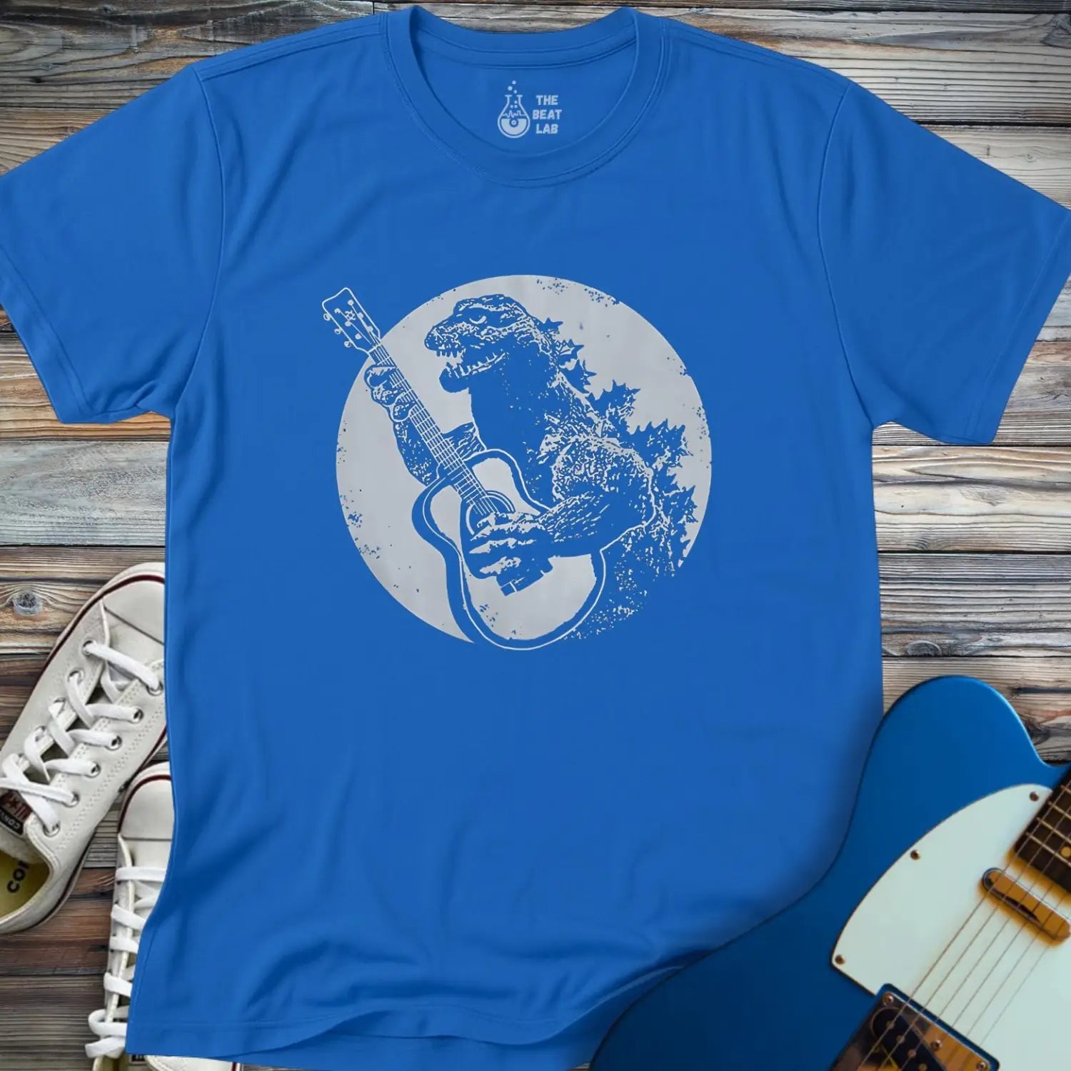 The Guitar Rex T-shirt features a blue tee with Godzilla strumming an acoustic guitar before a moonlit backdrop, styled on wood alongside a blue electric guitar and white sneakers.