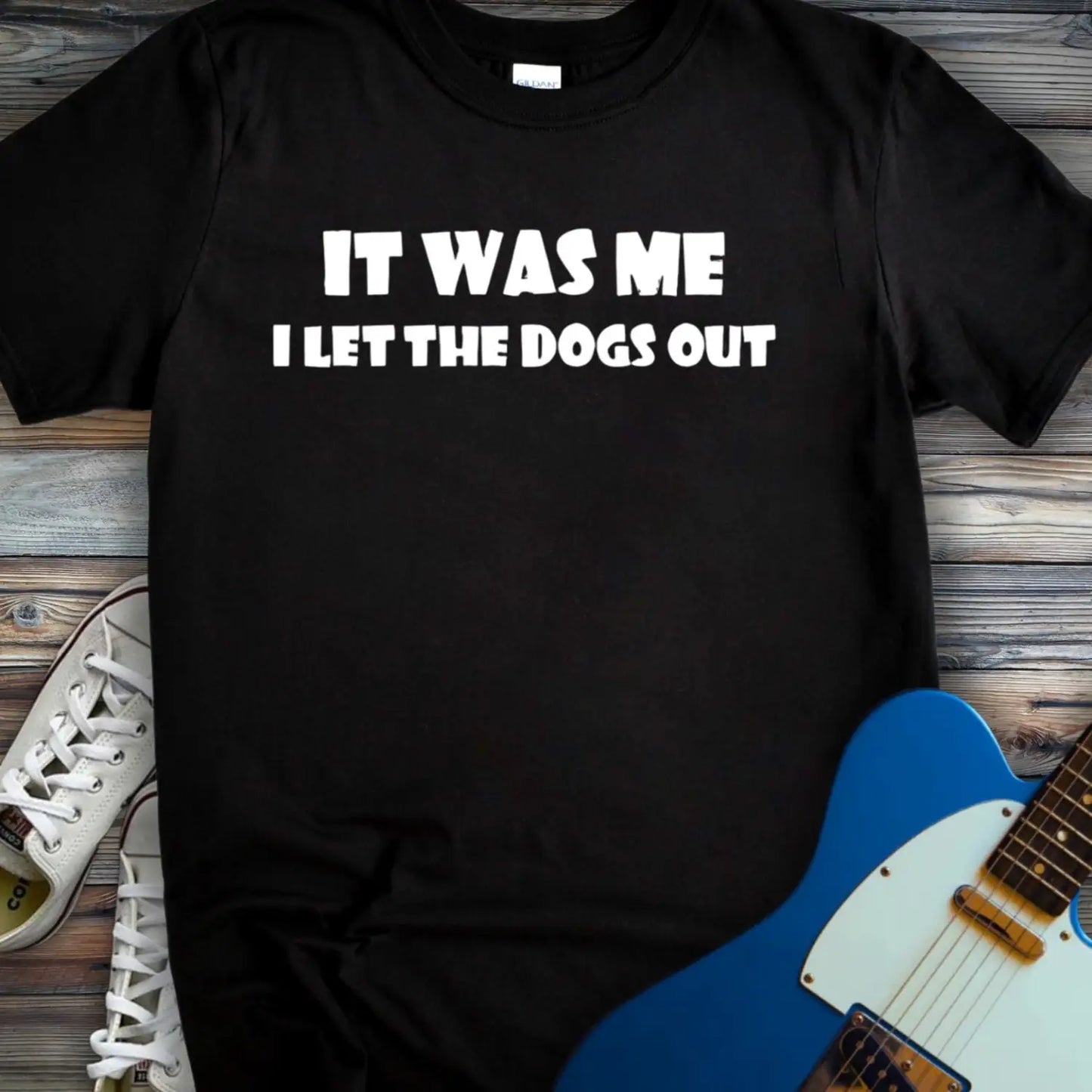 Black color funny graphic t-shirt with bold white text that says It Was Me I Let The Dogs Out, humorous music inspired meme tee for men and women, unisex casual streetwear shirt displayed flat lay with sneakers and electric guitar.