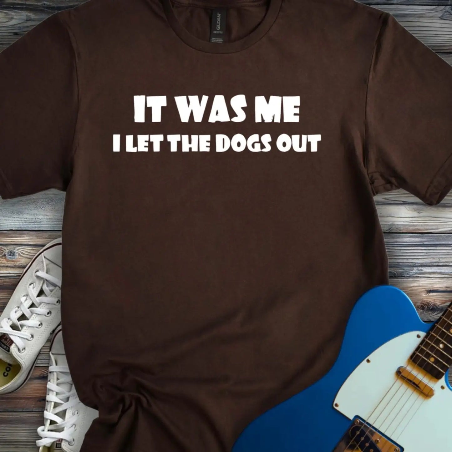 Dark Chocolate color funny graphic t-shirt with bold white text that says It Was Me I Let The Dogs Out, humorous music inspired meme tee for men and women, unisex casual streetwear shirt displayed flat lay with sneakers and electric guitar.