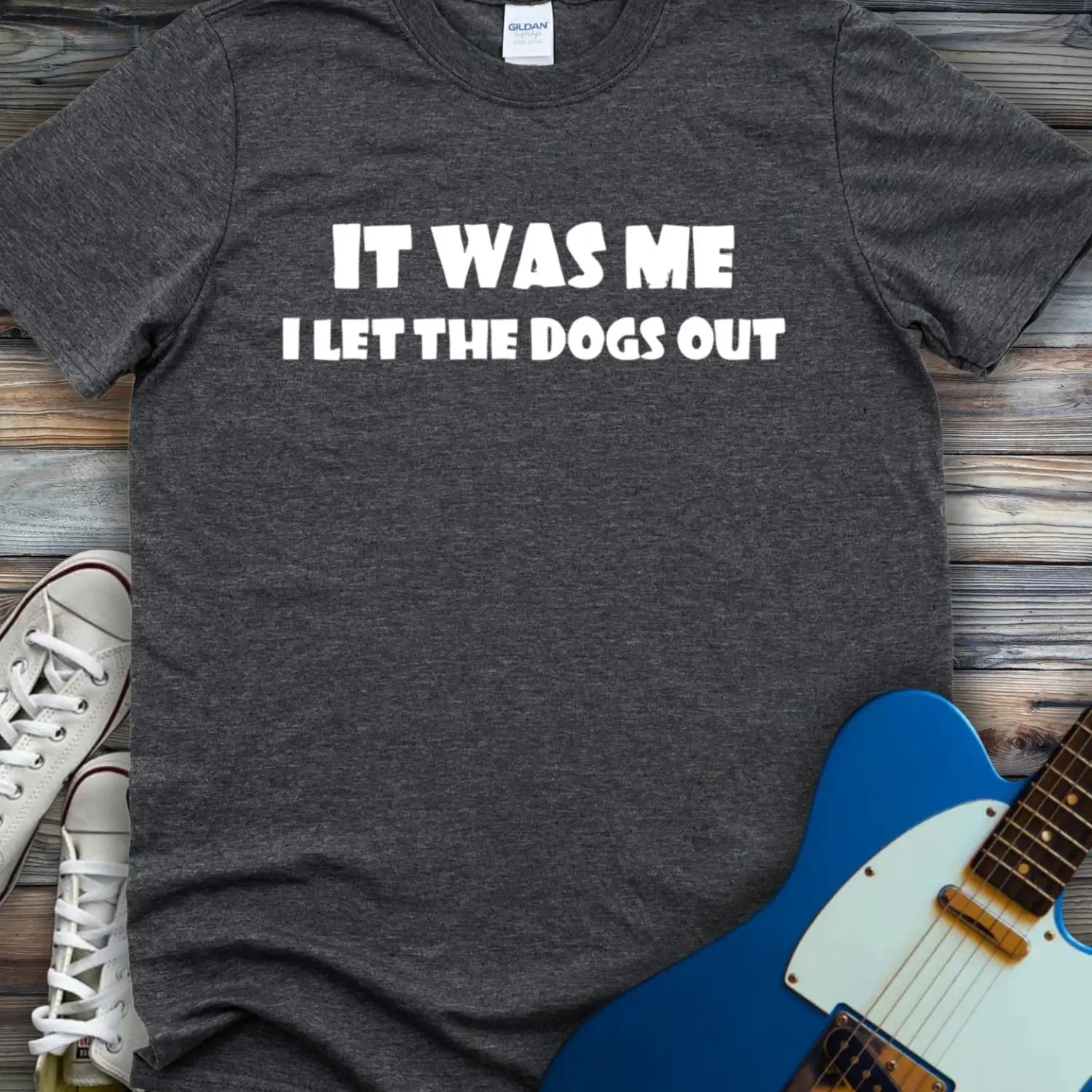 Dark Heather Grey color funny graphic t-shirt with bold white text that says It Was Me I Let The Dogs Out, humorous music inspired meme tee for men and women, unisex casual streetwear shirt displayed flat lay with sneakers and electric guitar.
