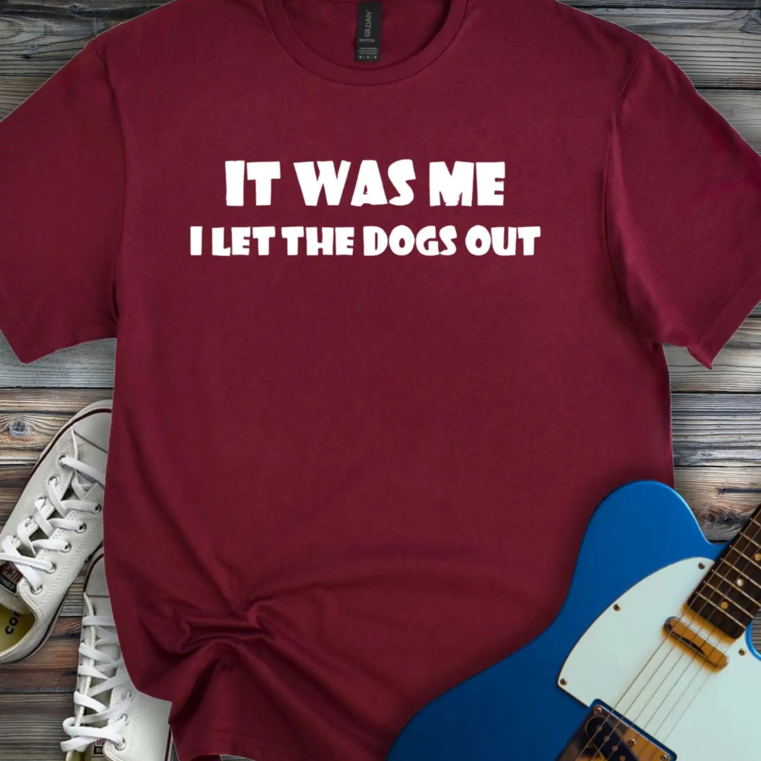 Maroon color funny graphic t-shirt with bold white text that says It Was Me I Let The Dogs Out, humorous music inspired meme tee for men and women, unisex casual streetwear shirt displayed flat lay with sneakers and electric guitar.