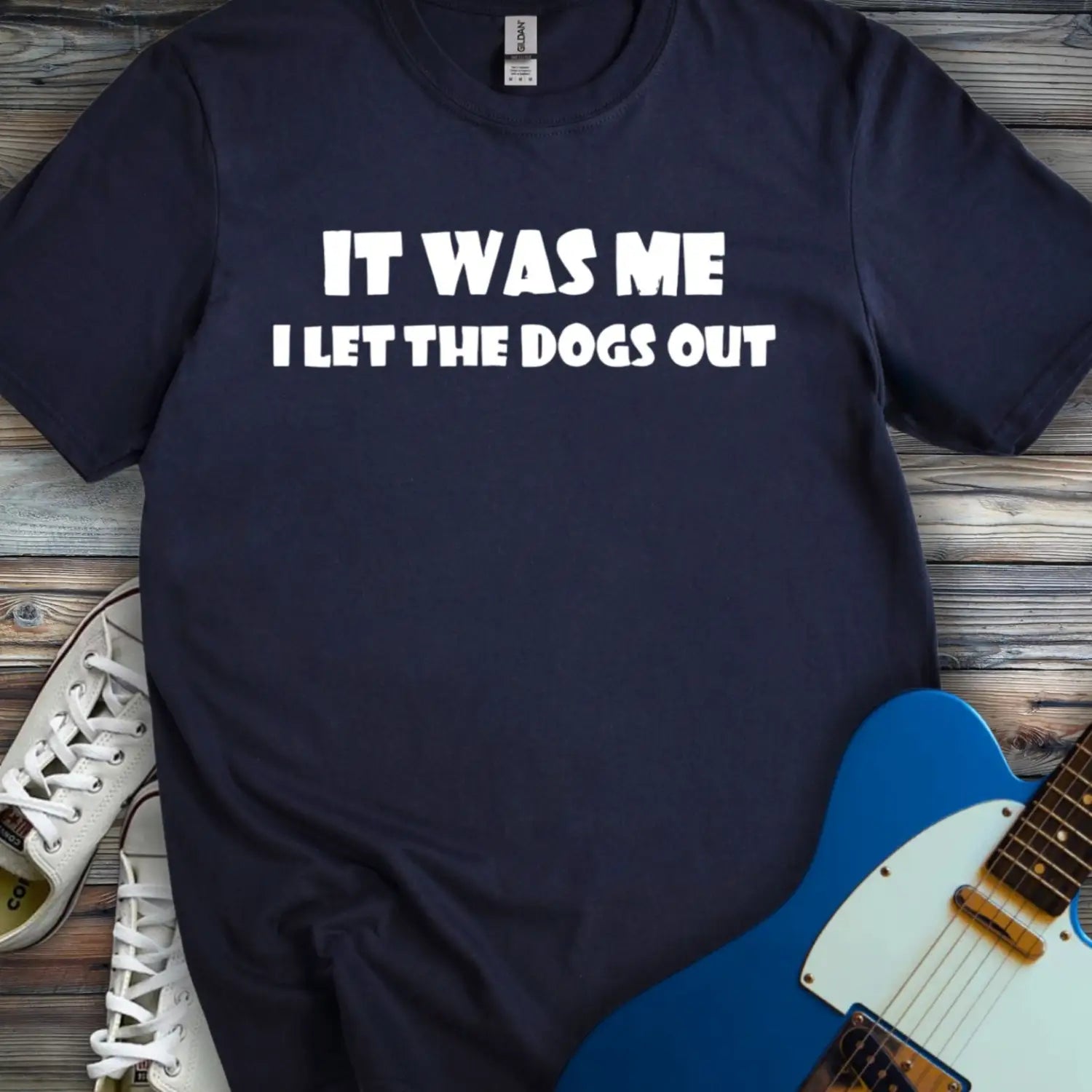 Navy blue color funny graphic t-shirt with bold white text that says It Was Me I Let The Dogs Out, humorous music inspired meme tee for men and women, unisex casual streetwear shirt displayed flat lay with sneakers and electric guitar.