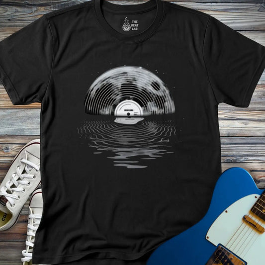 The Moon Vinyl T-shirt features a black design with a vinyl record forming a rising sun over rippling water, showcased on wood beside white sneakers and a blue electric guitar.