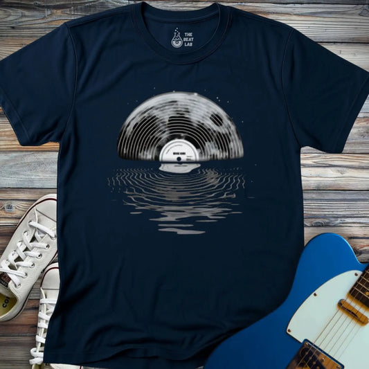 The Moon Vinyl T-shirt is a navy blue tee with a vinyl record half-submerged in water, creating a reflection. Pictured with white sneakers and a blue electric guitar on a wooden surface.