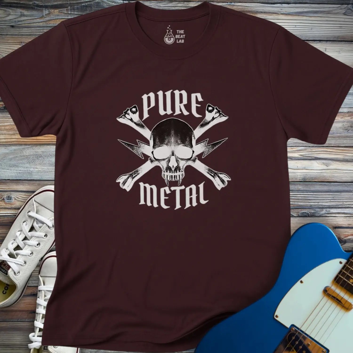 The Pure Metal T-shirt in maroon features a skull and crossed bones graphic with "PURE METAL" text. It’s displayed on a wooden surface beside white sneakers and a blue electric guitar.