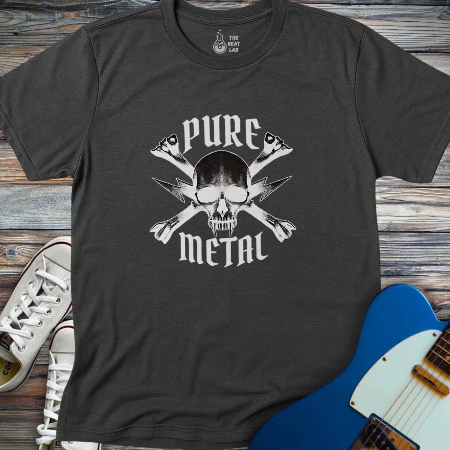 The Pure Metal T-shirt, dark gray with a skull and crossed lightning bolts and "PURE METAL" text, is displayed on a wooden surface alongside white sneakers and a blue electric guitar.