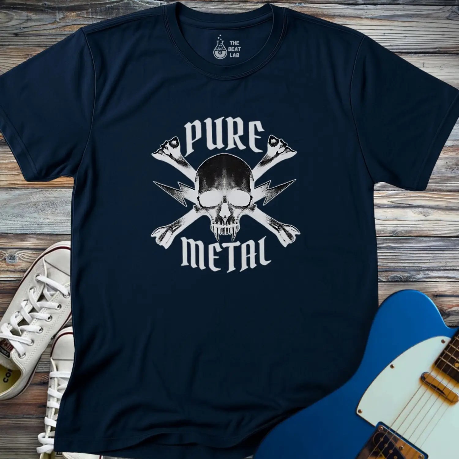 The Pure Metal T-shirt is a navy blue tee with a skull and crossbones graphic featuring crossed guitar necks and "PURE METAL" text. It's displayed on a wooden surface beside white sneakers and a blue electric guitar.