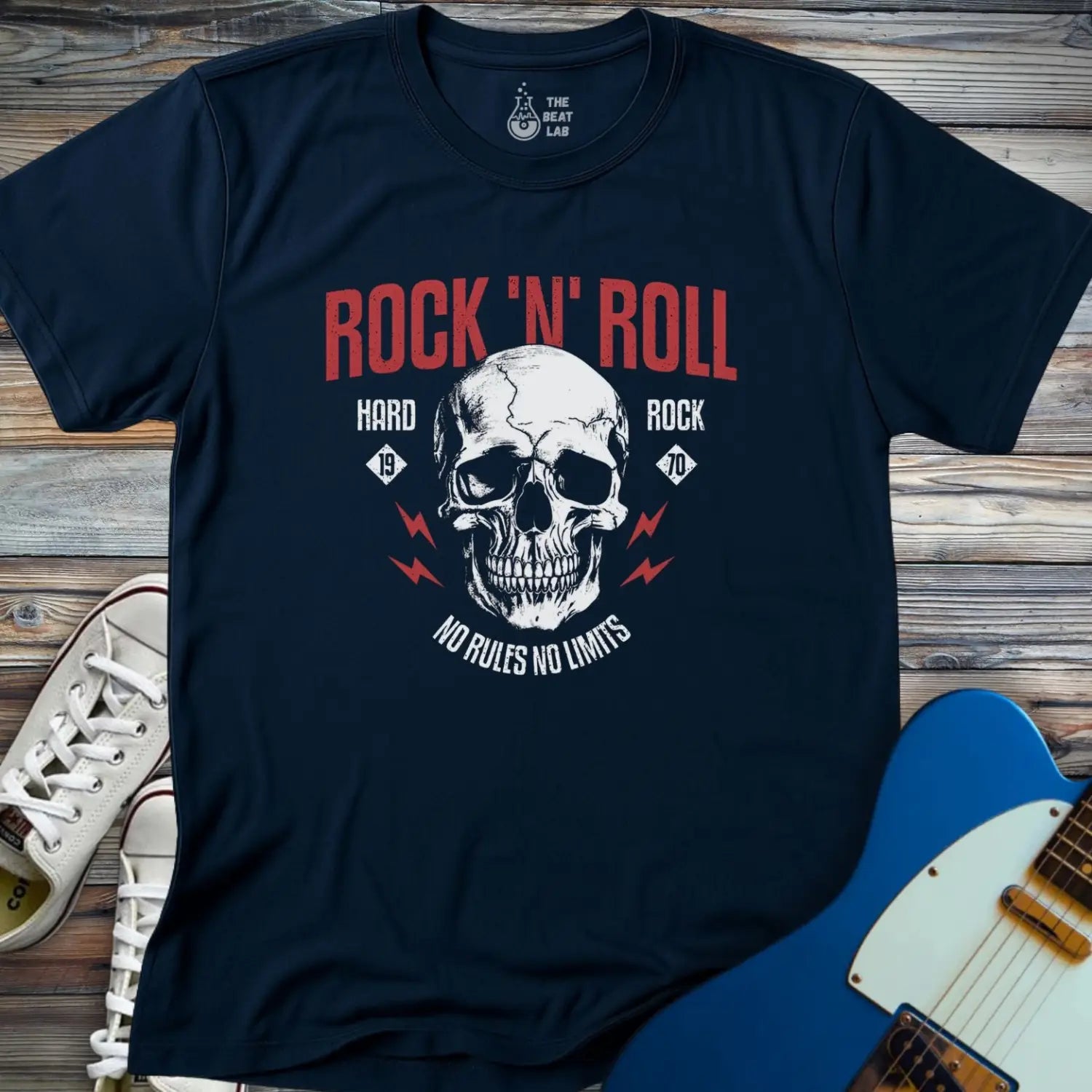 The Rock 'N' Roll 1970 T-shirt is a navy blue tee with a white skull, red lightning bolts, and “ROCK ‘N’ ROLL,” “HARD ROCK,” and “NO RULES NO LIMITS” text, shown beside white sneakers and a blue electric guitar on wood.